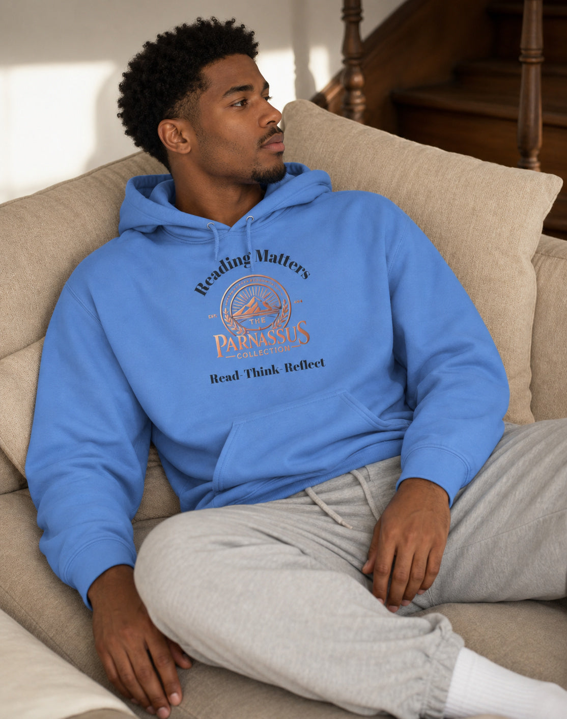 A man sitting on a beige couch wearing a Carolina blue pullover hoodie with a graphic print that reads 'Reading Matters' and above the image of the 'PARNASSUS COLLECTION' logo with the words 'Read Think Reflect' printed below.