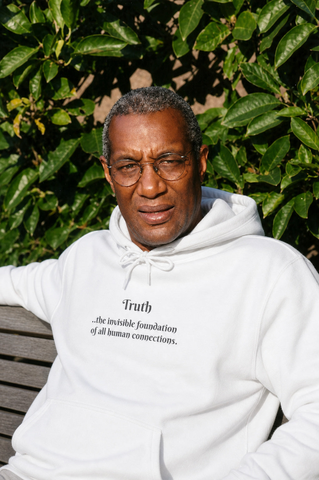 A man sitting on a park bench wearing a white pullover hoodie and matching drawstrings. Centered on the front just above a kangaroo pocket is the word "Truth" followed these words, "..the invisible foundation of all human connections."