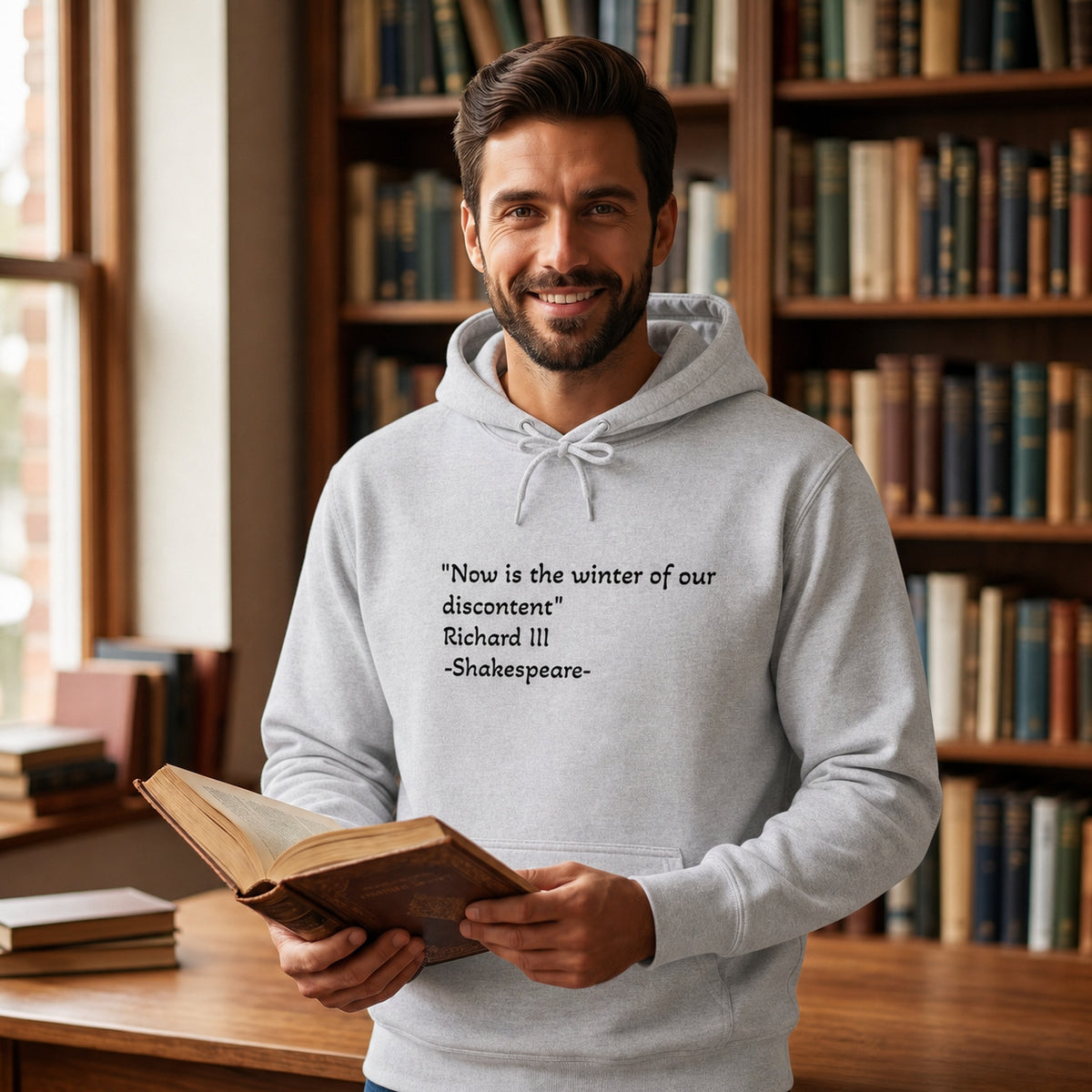A smiling man in a library wearing a graphite heather hoodie with this printed quote from Richard III, Shakespeare, "Now is the winter of our discontent."